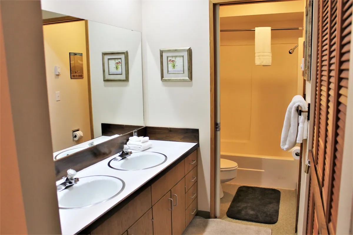 A bright, inviting bathroom featuring a double sink vanity, large mirrors, fresh towels, and framed artwork on the walls. The design is both functional and stylish, ideal for morning or evening routines.