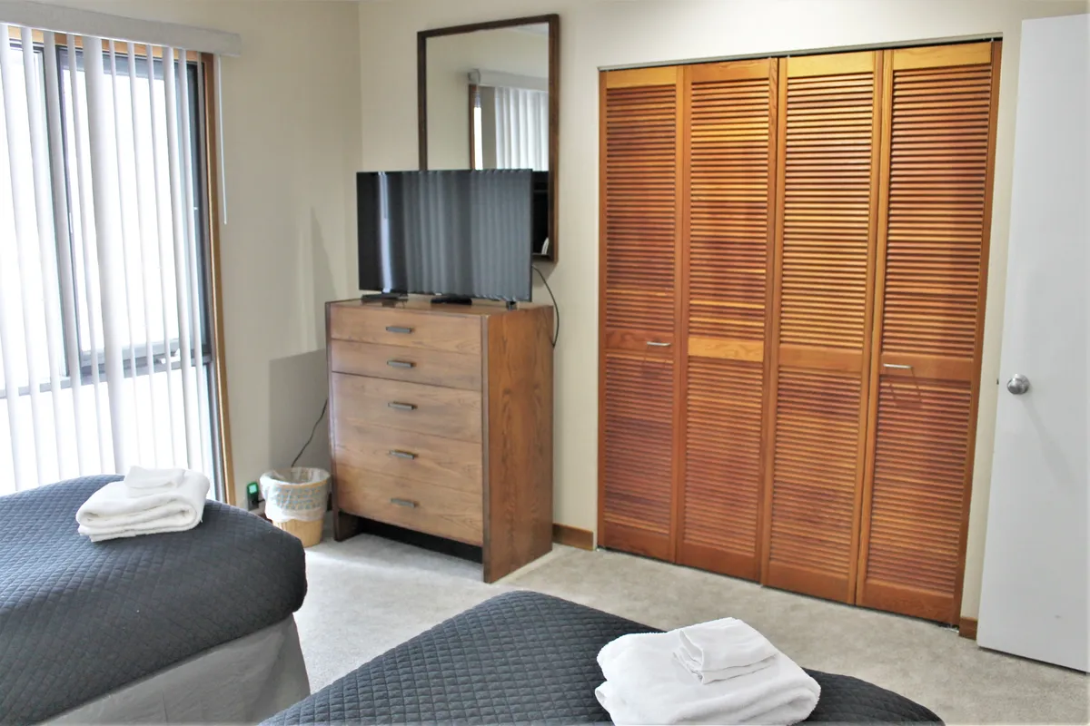 A practical and cozy second bedroom with two twin beds, each topped with neatly folded towels, a wooden dresser, a closet, and a television. The arrangement highlights the room’s flexibility for families or groups and its entertainment options.