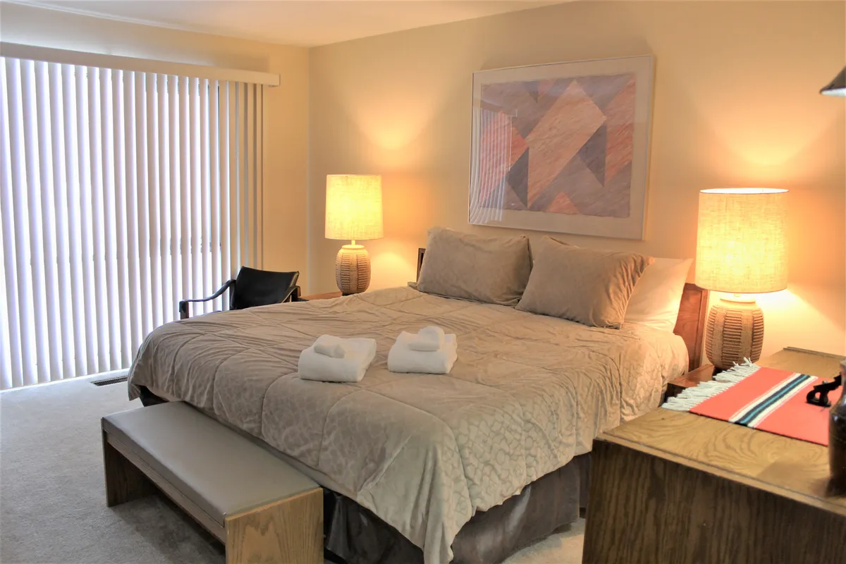 A serene primary bedroom featuring a plush king bed adorned with cozy textiles, flanked by stylish bedside tables and lamps, complemented by tasteful artwork and a bench at the foot of the bed. Soft lighting and calming decor create a welcoming, restful atmosphere.