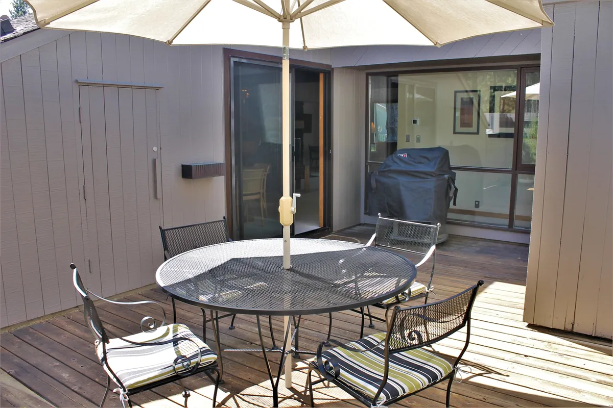 This image features a round table with an umbrella and four chairs set on a patio, positioned right outside sliding glass doors. A grill is visible nearby, and the scene is framed with casual patio decor, inviting guests to dine, relax, or socialize outdoors.