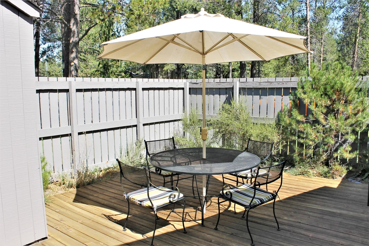 A tranquil wooden deck outfitted with a round table, black metal chairs, a large umbrella, and lush surrounding greenery. The setting feels peaceful and inviting, ideal for outdoor dining or relaxing while enjoying the scenery.