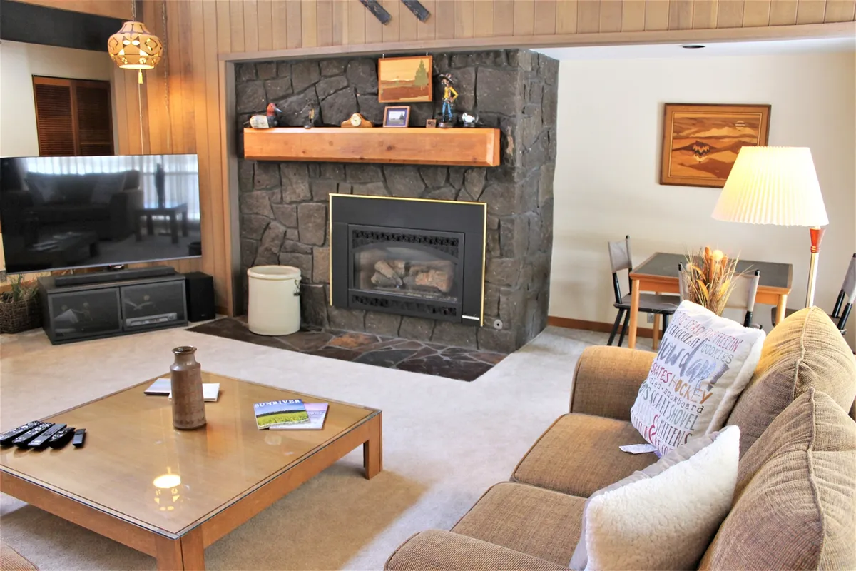 A cozy, open living space featuring a stone-faced fireplace with a wooden mantel, a comfortable sofa, a wooden coffee table, and a large television. The area is styled with warm accents and offers both seating and entertainment options, set within a spacious layout that connects to the rest of the living area.