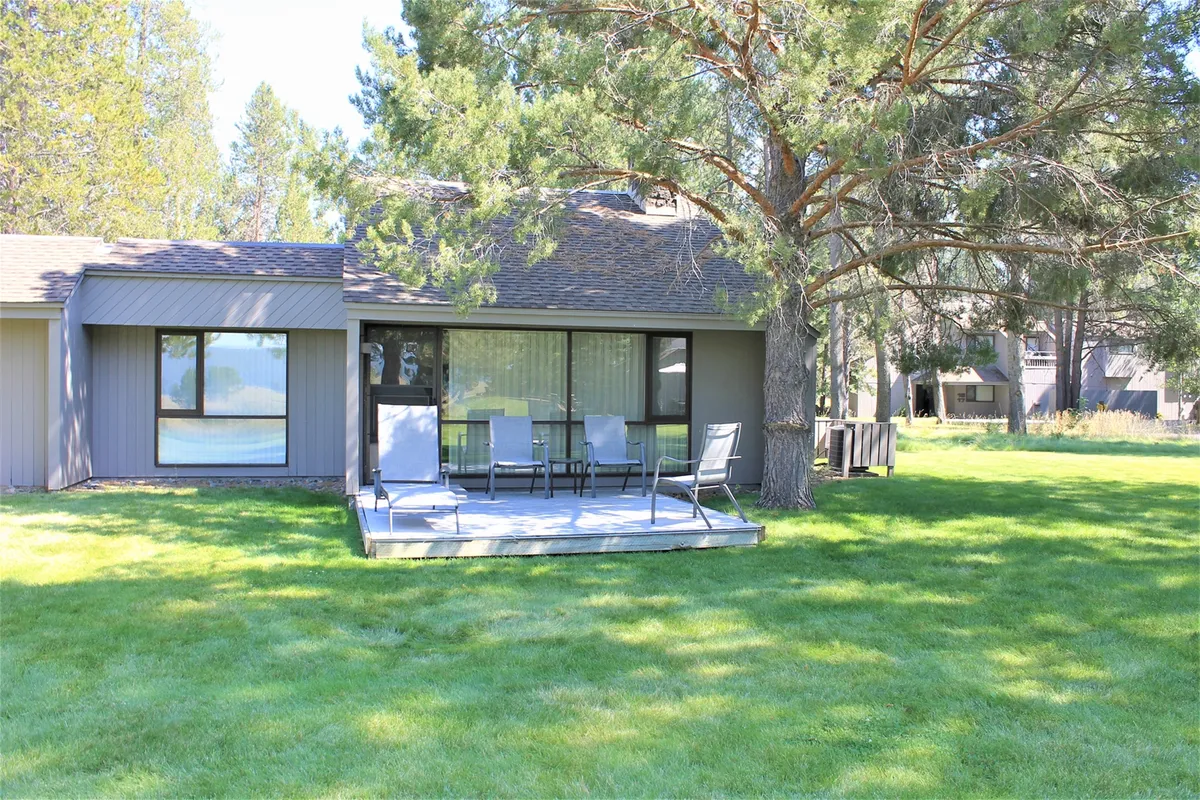 The image showcases a spacious deck area nestled beneath a large tree with the house’s windows in view. The deck invites outdoor relaxation, while the mature landscaping and open sky suggest a peaceful connection to nature just outside the door.